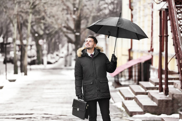 Young man in warm clothes with dark umbrella and briefcase outdoors - Фото, изображение