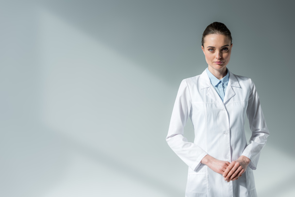 médico fêmea grave em casaco branco olhando para a câmera em cinza
 - Foto, Imagem