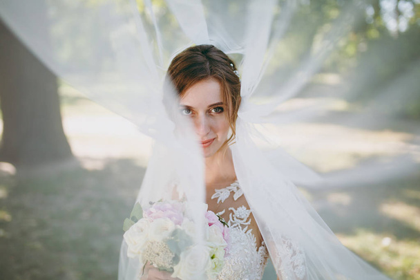 Beautiful wedding photosession. Portrait of the young smiling bride in a white lace dress and bouquet of white and pink flowers in a fluttering veil in a large green garden on weathery sunny day - Fotoğraf, Görsel