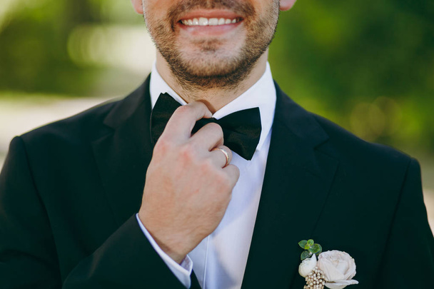 Прекрасная свадебная фотосессия. Улыбающийся жених в черном костюме, белая рубашка с бутоньеркой и кольцо на пальце выпрямляет бабочку на расплывчатом зеленом фоне сада
 - Фото, изображение
