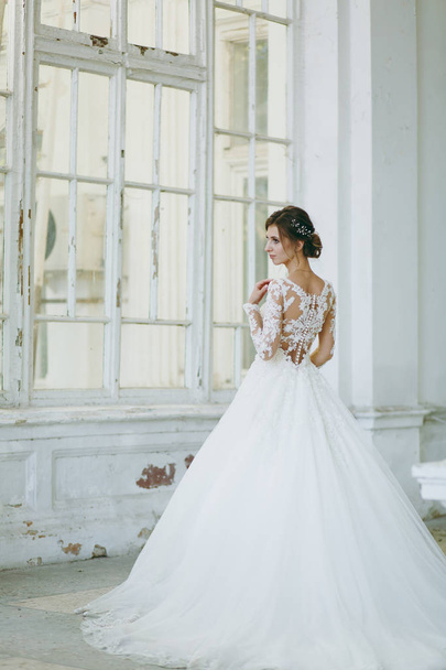 Beautiful wedding photosession. Young bride in a white lace dress with a long plume with an exquisite hairstyle in vintage interior on the veranda of an old house with columns near the garden - Foto, afbeelding