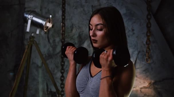 Sexy sporty girl in short shorts and inflated press with dumbbells indoors on the background of a stone gray wall raises her hands with dumbbells for bicep training. Close up - Footage, Video
