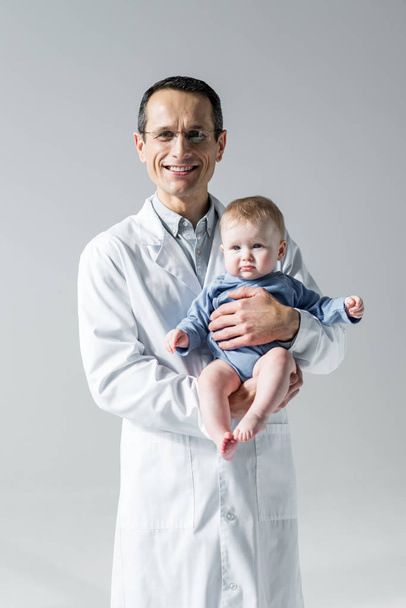 pediatra adulto sonriente sosteniendo al pequeño bebé y mirando a la cámara aislada en gris
 - Foto, imagen