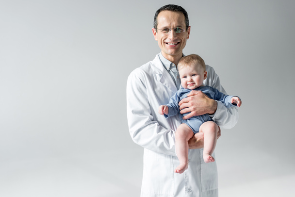 glücklicher erwachsener Kinderarzt mit kleinem Baby und Blick in die Kamera isoliert auf grau - Foto, Bild