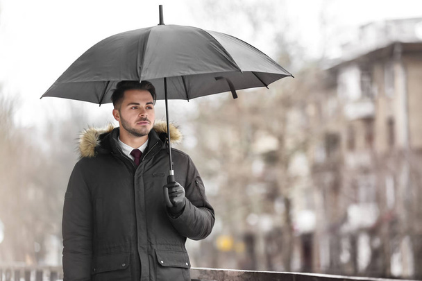 Young man in warm clothes with dark umbrella outdoors - Fotó, kép