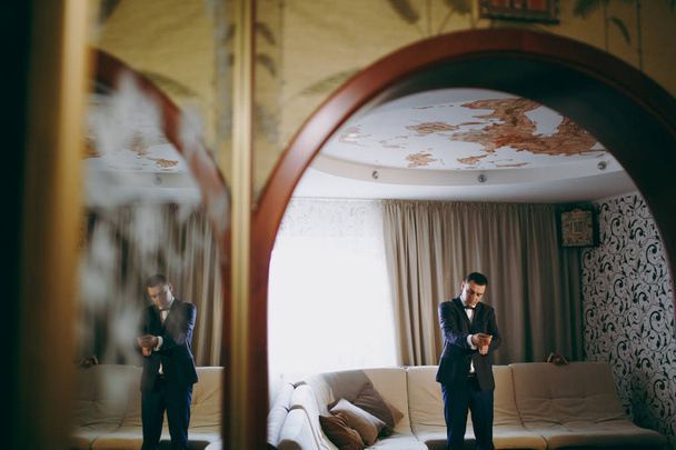Preparação matinal dos recém-casados para a cerimônia de casamento. O noivo na camisa branca, calças azuis, jaqueta e gravata borboleta põe abotoaduras em mangas no quarto com papel de parede modelado. Casamento desgaste
 - Foto, Imagem