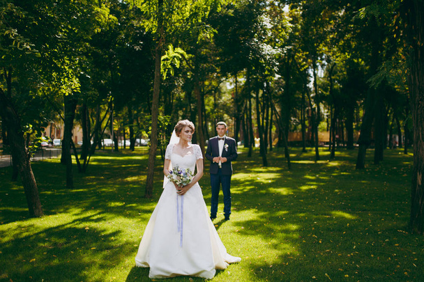 Güzel düğün photosession. Mavi resmi elbise ve beyaz elbise ve peçe kokulu güzel hairdress güneşli bir büyük yeşil parkta bir yürüyüş ile onun zarif gelin yakışıklı damat - Fotoğraf, Görsel