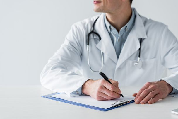 Schnappschuss eines Arztes, der am Arbeitsplatz sitzt und Diagnose in Klemmbrett schreibt, isoliert auf weiß - Foto, Bild