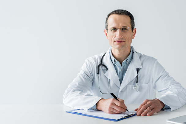 handsome adult doctor sitting at workplace and writing diagnosis in clipboard isolated on white - Photo, Image
