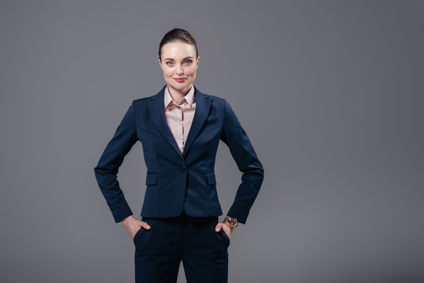 smiling adult businesswoman with hands in pockets looking at camera isolated on grey - Foto, Bild