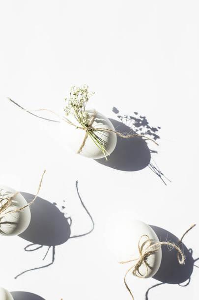 White minimalistic Easter eggs decorated with twine and flowers of gypsophila on a white background in bright light with deep shadows. Easter concept. Top view, flat lay - Fotoğraf, Görsel