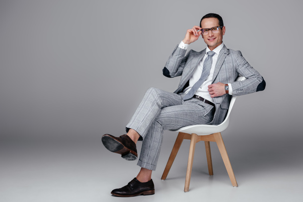 smiling adult businessman in stylish suit sitting on chair on grey - Fotó, kép