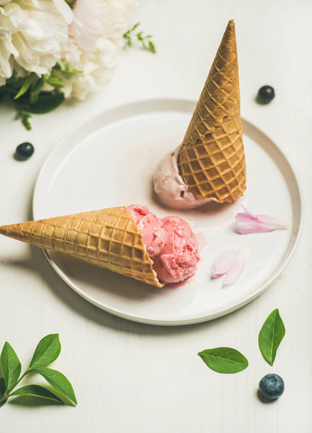 Pink strawberry and coconut ice cream scoops, sweet cones and peony flowers bouquet on plate over white background - Фото, изображение