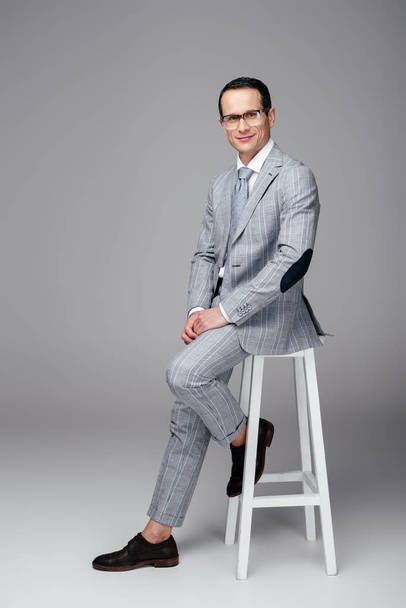 handsome adult businessman in stylish suit sitting on chair on grey - Foto, immagini