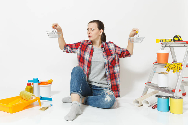 Shocked upset sad woman holds metal grocery baskets for shopping. Instruments for renovation apartment isolated on white background. Wallpaper, accessories, tools for painting. Repair home concept. - Photo, Image