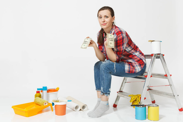 Upset female holds tight of dollars, cash few money, sits on ladder with instruments for renovation apartment isolated on white background. Wallpaper, gluing accessories painting tools. Repair concept - Photo, Image