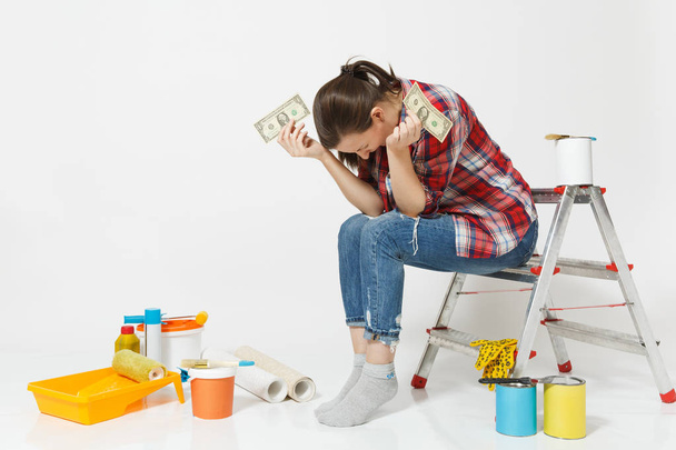 Järkyttynyt nainen omistaa tiukka dollareita, käteistä vähän rahaa, istuu tikkailla välineitä remontin huoneisto eristetty valkoisella taustalla. Taustakuva, liimaus tarvikkeet maalaus työkaluja. Korjauskäsite
 - Valokuva, kuva