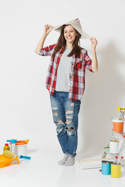 Full length portrait of woman in newspaper hat standing near instruments for renovation apartment home isolated on white background. Wallpaper, accessories for gluing, painting tools. Repair concept. - Photo, Image
