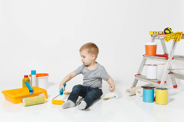 Little cute boy sitting on floor with instruments for renovation apartment room isolated on white background. Wallpaper, gluing accessories, painting tools. Repair home. Parenthood, childhood concept. - Foto, afbeelding