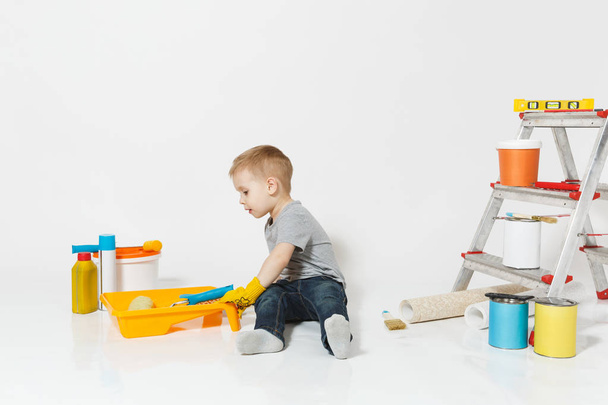 Little cute boy in yellow gloves with instruments for renovation apartment room isolated on white background. Wallpaper, gluing accessories, painting tools. Repair home. Parenthood, childhood concept. - Photo, Image