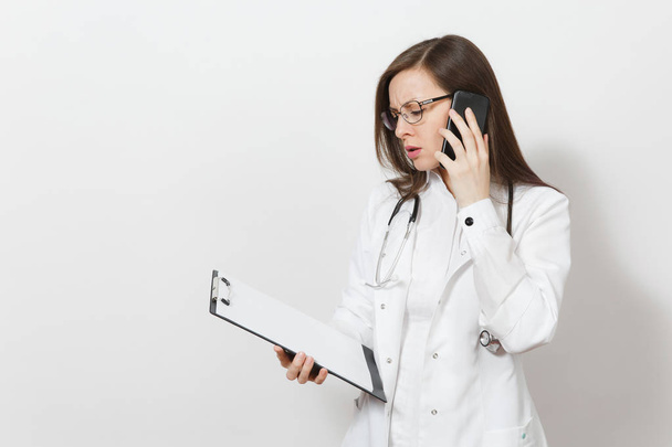 Ciddi genç doktor stetoskop, beyaz arka plan üzerinde izole gözlük kadınla. Kadın doktor tıbbi elbise cep telefonu kullanıyor musun, sağlık kartı not defteri klasör üzerinde tutun. Sağlık personeli kavramı. - Fotoğraf, Görsel