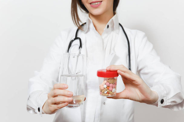 Κοντινό πλάνο περικοπεί shot χαμογελαστό γυναίκα γιατρός με το στηθοσκόπιο που απομονώνονται σε λευκό φόντο. Γυναίκα γιατρό σε ιατρική φόρεμα κάτοχος μπουκάλι με χάπια, ποτήρι νερό. Νοσηλευτικού προσωπικού, ιατρική έννοια. - Φωτογραφία, εικόνα