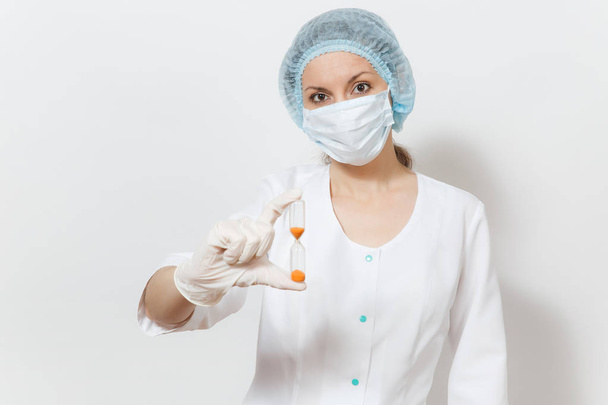 Young doctor woman with face mask, sterile hat, gloves isolated on white background. Female surgeon doctor in medical gown holds hourglass. Healthcare personnel, medicine concept. Time is running out. - Φωτογραφία, εικόνα