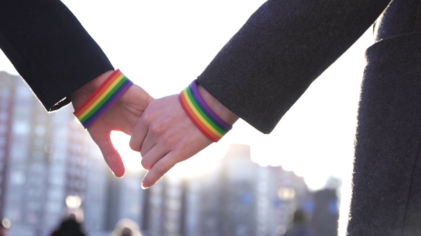 Two hands together with LGBT bracelets against the background of the street and the sun - Footage, Video
