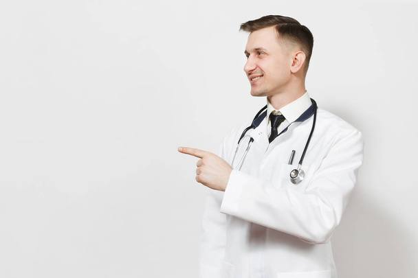 Smiling handsome young doctor man isolated on white background. Male doctor in medical gown, stethoscope pointing index finger aside on copy space. Healthcare personnel, health, medicine concept. - Foto, Bild