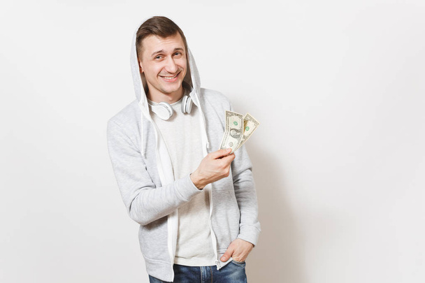 Young handsome smiling man student in t-shirt, blue jeans and light sweatshirt with hood with headphones holds two one-dollar bills and rejoiced isolated on white background. Concept of emotions - Photo, Image