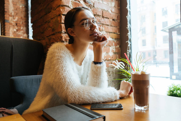 beautiful young girl in a white sweater and glasses sits in a cafe and looks thoughtfully out the window - Фото, изображение