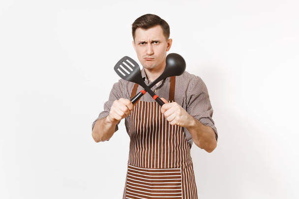Young warlike man chef or waiter in striped brown apron, shirt holding black ladle or kitchen spoon, spatula isolated on white background. Male housekeeper or houseworker. Kitchenware, cuisine concept - Photo, Image