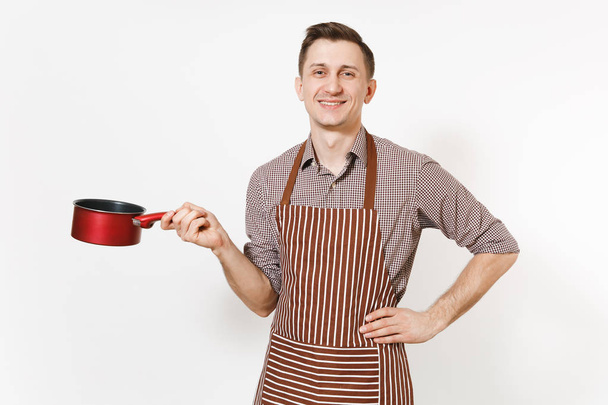 Young smiling man chef or waiter in striped brown apron, shirt holding red empty stewpan, pan or pot isolated on white background. Male housekeeper or houseworker. Kitchenware and cuisine concept. - Photo, Image
