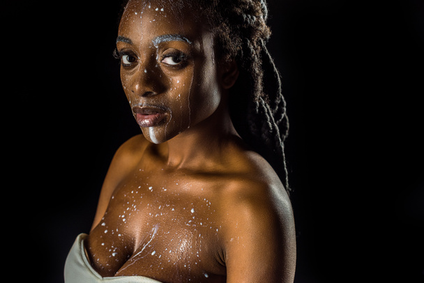 sensual joven afroamericana mujer con leche derramada en la cara mirando a la cámara aislada en negro
 - Foto, Imagen