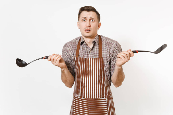 Bewildered man chef or waiter in striped brown apron, shirt holding black ladle or kitchen spoon, spatula isolated on white background. Male housekeeper or houseworker. Kitchenware, cuisine concept. - Valokuva, kuva