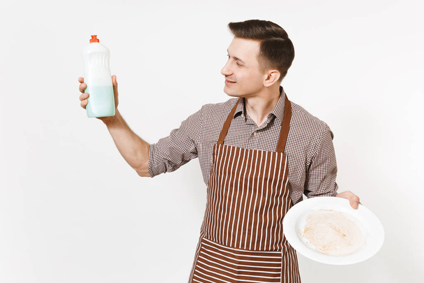 Man in striped brown apron, shirt holds white round dirty plate, detergent. Washing dishes with cleaner liquid in bottle isolated on white background. Male housekeeper, houseworker or domestic worker. - Fotografie, Obrázek