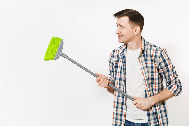 Young strange fun housekeeper man in checkered shirt holding and sweeping with green broom isolated on white background. Male doing house chores. Copy space for advertisement. Cleanliness concept. - Фото, зображення