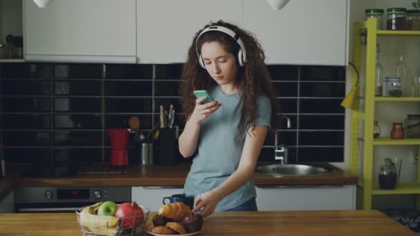 curly caucasian pretty teenage girl with headphones taking photo of breakfast, moving cup, taking cutting board, put red pomegranate on it and taking another photo - Materiał filmowy, wideo