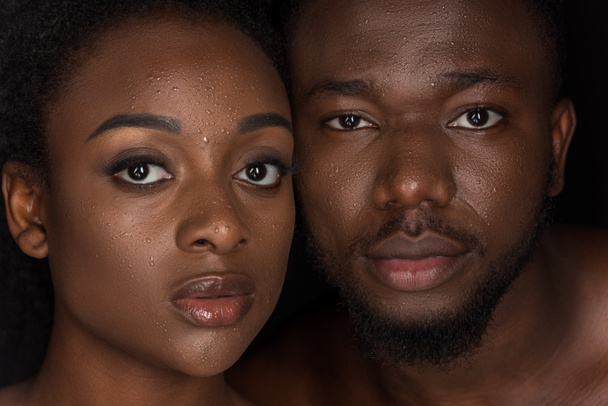 young african american couple with water drops on faces looking at camera on black - Фото, изображение