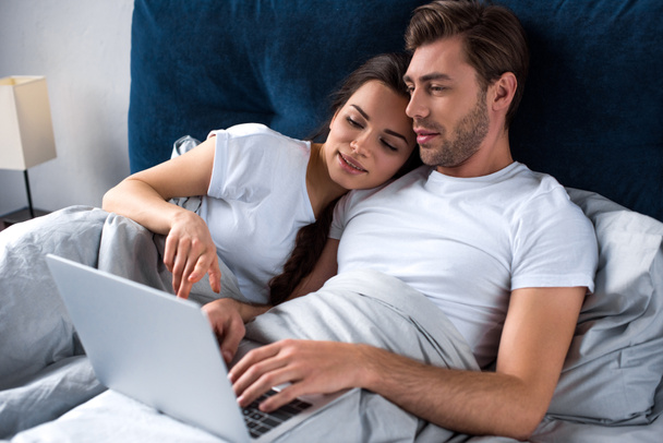 Young man and woman working on laptop while lying in bed - Fotoğraf, Görsel