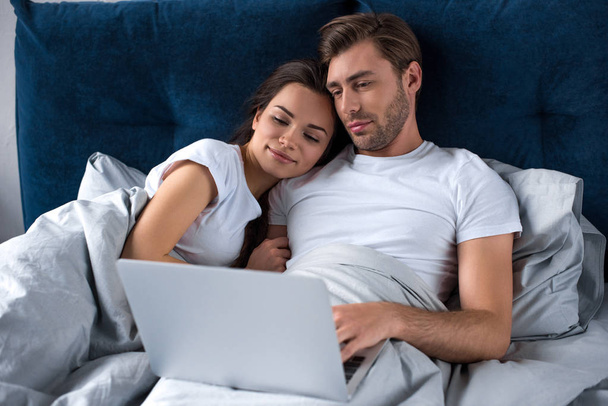 Smiling couple working on laptop while lying in bed - 写真・画像