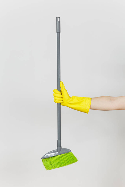 Κοντινό πλάνο της γυναικείας χέρι σε κίτρινα γάντια κατέχει μεγάλη γκρι σκούπα με πράσινο λαχνών για τον καθαρισμό και σαρωτικές απομονώνονται σε λευκό φόντο. Καθαρίζοντας προμήθειες έννοια. Αντίγραφο χώρος για διαφήμιση - Φωτογραφία, εικόνα
