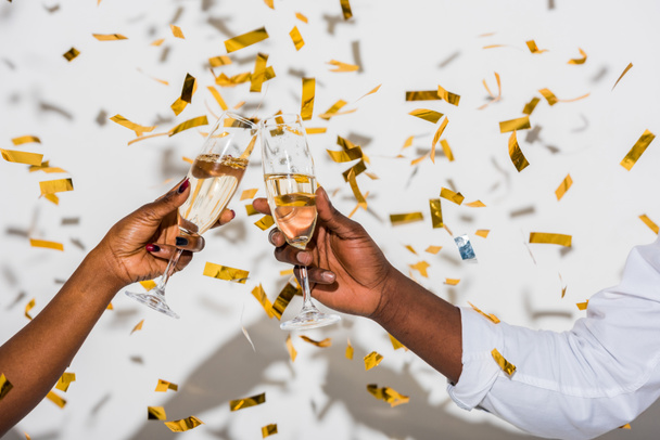 Close-up deelaanzicht Afrikaanse Amerikaanse echtpaar rammelende glazen champagne op wit met gouden confetti - Foto, afbeelding