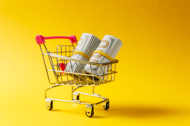 Supermarket grocery push cart for shopping with pink elements on handle with bundles of dollars money banknote cash isolated on yellow background. Concept of shopping. Copy space for advertisement. - Photo, Image