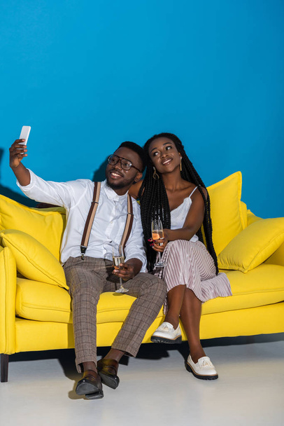 happy stylish young african american couple holding glasses of wine and taking selfie with smartphone on sofa  - Foto, immagini
