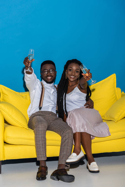 Gelukkig stijlvolle Afrikaanse Amerikaanse echtpaar drinken champagne en lachend op camera zittend op de Bank - Foto, afbeelding