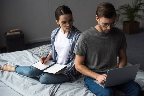 Woman writing in notepad and looking at man working on laptop while sitting on bed - Photo, Image