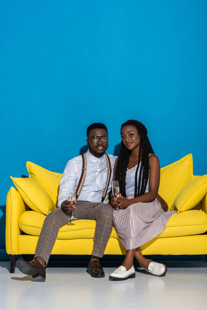 young african american couple holding glasses of wine and smiling at camera while sitting on yellow sofa  - Fotoğraf, Görsel