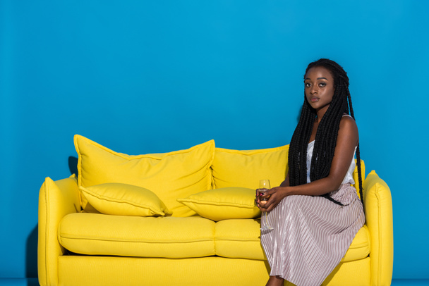 beautiful young african american woman holding glass of wine while sitting on sofa and looking at camera on blue - Photo, Image