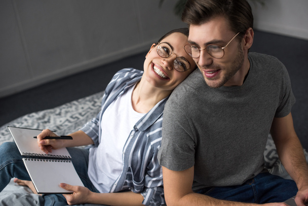 Happy man and woman in glasses writing in notepad while lying in bed - Photo, Image